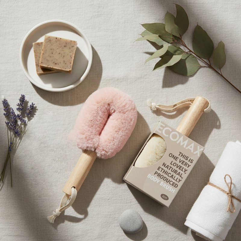 Pink body brush with wooden handle, soap, and towel on a neutral background