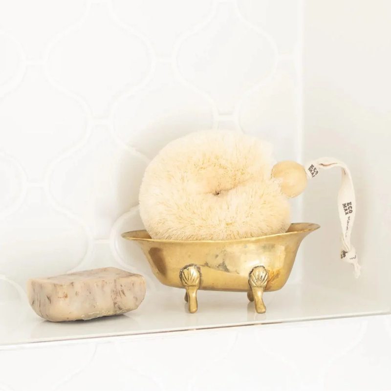 Natural spa brush in a gold dish, and soap on a white surface
