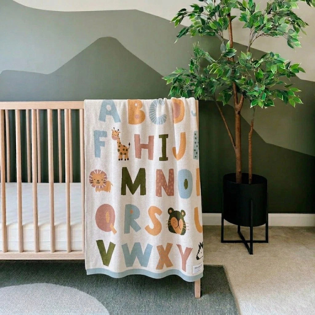 Children's alphabet blanket hanging over a crib in a room with a plant.