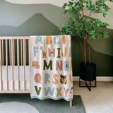 Children's alphabet blanket hanging over a crib in a room with a plant.