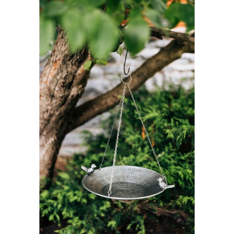Metal bird bath hanging from a tree branch with green foliage in the background