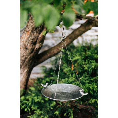 Metal bird bath hanging from a tree branch with green foliage in the background