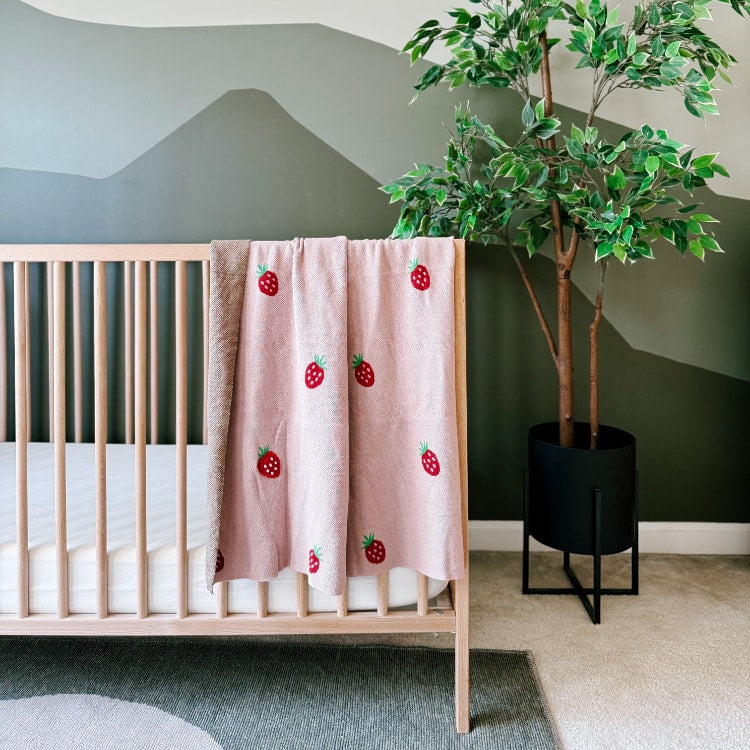 Nursery room with a crib, pink baby blanket with red strawberry, and a potted plant against a gray wall.