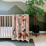 Crib with a patterned blanket featuring woodland animals and trees, against a green wall with a plant.