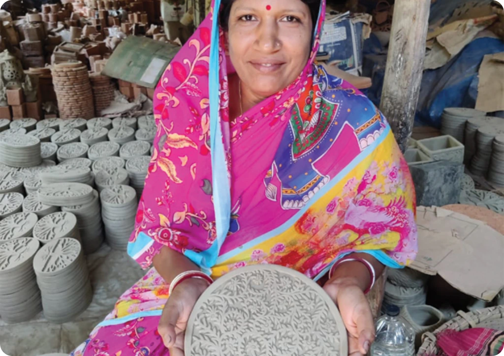Woman in a colorful saree holding a clay disc in an outdoor setting with pottery items around.